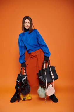 brunette woman in autumnal clothes holding different bags on orange backdrop, black friday concept clipart