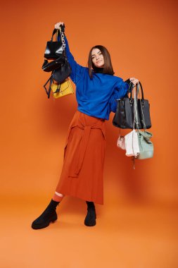 happy woman in autumnal clothes holding different handbags on orange backdrop, black friday concept clipart