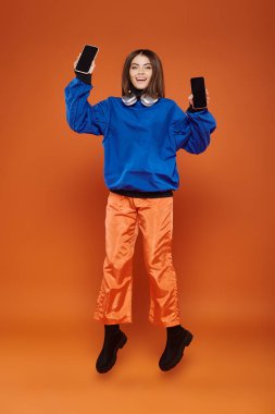 Moda kıyafetleri içinde neşeli bir kadın turuncu, siber pazartesi konseptiyle akıllı telefonlarla zıplıyor.