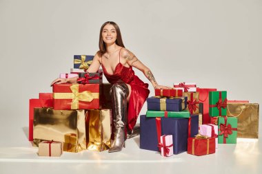 attractive young woman in red dress surrounded by presents looking at camera, holiday gifts concept clipart