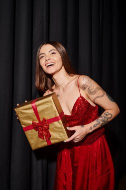 happy woman with tattoos and piercing smiling with black curtains on backdrop, holiday gifts concept clipart