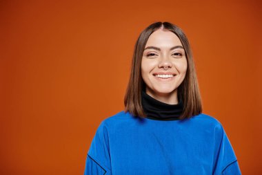 attractive smiley woman in casual blue jacket smiling cheerfully at camera on orange background clipart