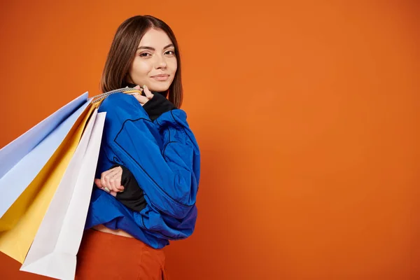 stock image positive woman in stylish autumnal clothes holding shopping bags on black friday, orange backdrop