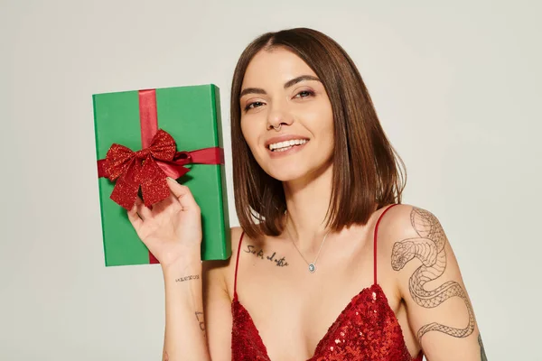 stock image portrait of cheerful woman with tattoos holding present on ecru backdrop, holiday gifts concept