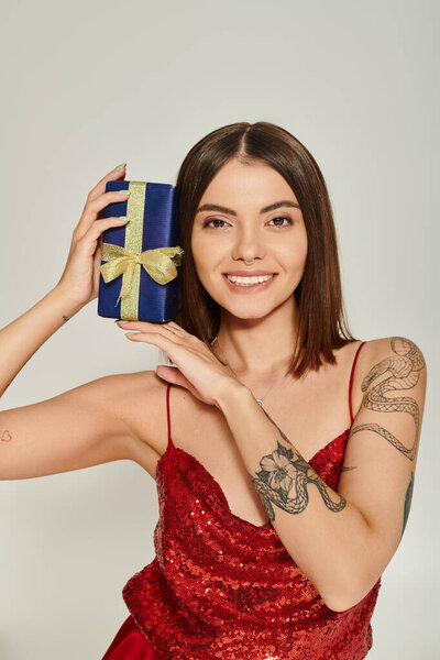 cheerful lady with tattoos and piercing holding present near face smiling at camera, holiday gifts