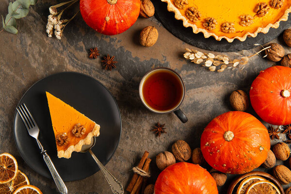 thanksgiving  pumpkin pie on black plate near warm tea and orange pumpkins on decorated stone table
