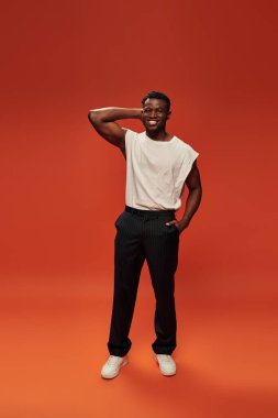 joyful african american man in white tank top with hand in pocket of black pants on red, full length clipart