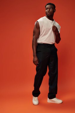 athletic african american man in black pants and white tank top looking away on red, full length clipart