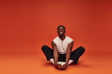 overjoyed african American man in white tank top and black pants sitting on red and orange backdrop clipart