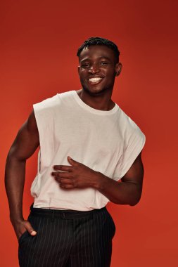 Siyah pantolon cebinde, kırmızı fonda kameraya bakan neşeli Afro-Amerikalı adam.