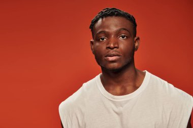 Beyaz kolsuz tişörtlü Afro-Amerikan genç bir adamın portresi. Kırmızı arka planda kameraya bakıyor.