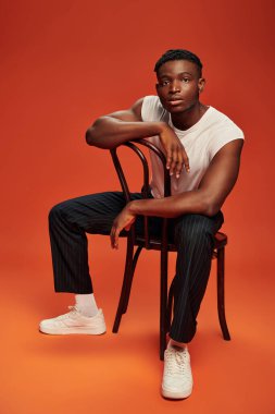 Beyaz kolsuz tişörtlü Afro-Amerikan bir adam sandalyede oturuyor ve kırmızı kameraya bakıyor.