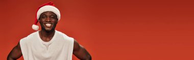 joyful african american guy in santa hat and white tank top looking at camera on red, banner clipart