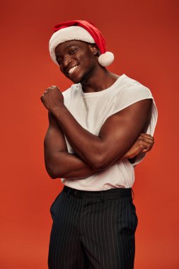 Ateşli ve atletik Afrikalı Amerikalı Noel Baba şapkalı ve beyaz atletli adam kırmızıda kameraya gülümsüyor.
