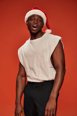 Noel Baba şapkalı ve beyaz tanklı Afro-Amerikalı adam kırmızı arka planda kameraya bakıyor.