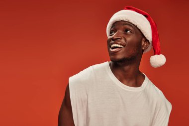 Şaşırmış ve çok sevinmiş, Noel Baba şapkalı Afrikalı Amerikalı adam gülümsüyor ve kırmızı arka plana bakıyor.