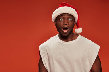 Şok olmuş Afrikalı Amerikalı Noel Baba şapkalı adam kırmızı arka planda ağzı açık kameraya bakıyor.