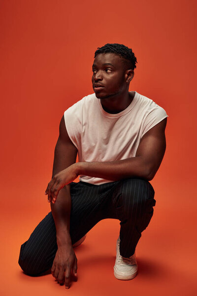 serious african american man in white tank top sitting on haunches and looking away on red backdrop