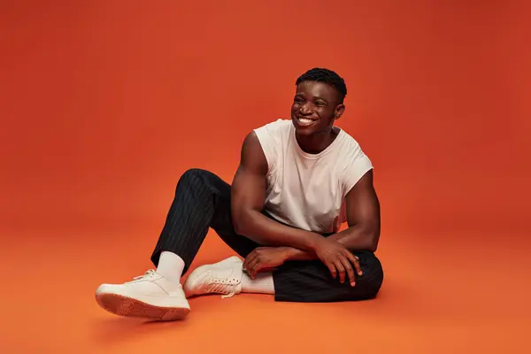happy african american man in stylish street wear sitting on red and orange backdrop, full length