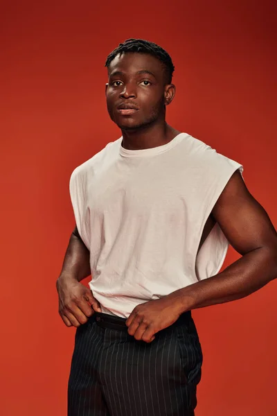 stock image pensive african american man in white tank top and black pants looking away on red backdrop