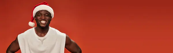 stock image joyful african american guy in santa hat and white tank top looking at camera on red, banner