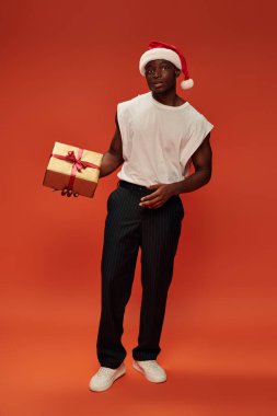 african american man in trendy casual clothes and santa hat with golden gift box on red, full length clipart