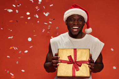 cheerful african american guy in christmas hat posing with gift boxes under falling confetti on red clipart