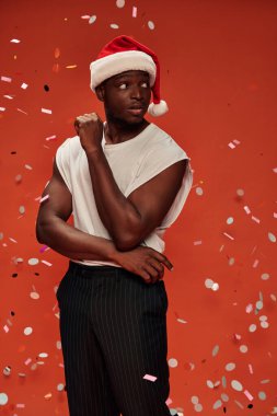 Beyaz kolsuz tişörtlü ve Noel Baba şapkalı genç Afro-Amerikan adam kırmızı konfeti altında başını çeviriyor.