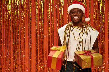 Noel Baba şapkalı mutlu Afro-Amerikalı adam, turuncu arka planda altın renkli süslemelerin yanında hediye kutularıyla.