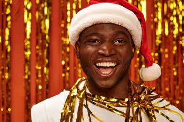 portrait of excited african american guy in santa hat laughing near golden tinsel on red backdrop clipart