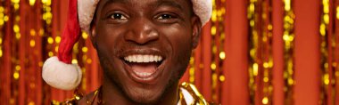 portrait of laughing african american guy in santa near shiny golden tinsel on red backdrop, banner clipart