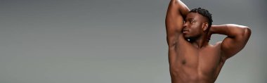 Kaslı ve üstsüz Afro-Amerikan erkek model. Gri pankartın arkasında elleri ile poz veriyor.