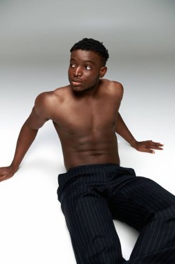 young shirtless african american man with muscular torso sitting and looking away on grey backdrop clipart