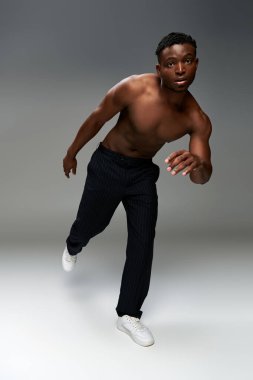 Üstsüz ve sportif Afro-Amerikalı siyah pantolonlu adam gri zeminde tek ayak üzerinde poz veriyor.