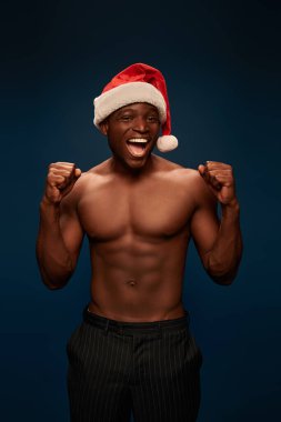 Noel Baba şapkalı Afrikalı Amerikalı sporcu lacivert arka planda zafer jesti sergiliyor.