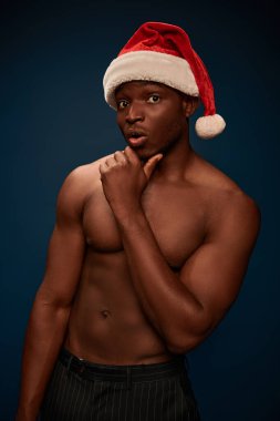 surprised african american man in santa hat with shirtless body looking at camera on dark blue clipart