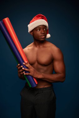 shirtless african american guy in santa hat holding colorful paper rolls on dark blue backdrop clipart