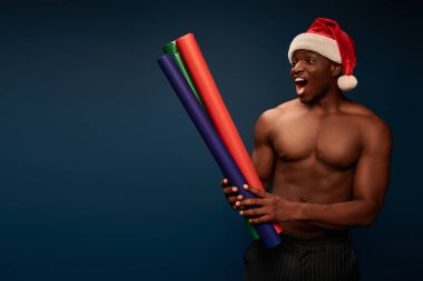 Çok neşeli atletik Afrikalı Amerikalı Noel Baba şapkalı adam elinde koyu renk duvar kağıtları tutuyor.