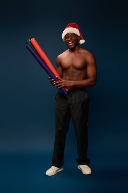 Neşeli kaslı Afrikalı Amerikalı Noel Baba şapkalı, lacivert arka planda renkli duvar kağıtları olan adam.