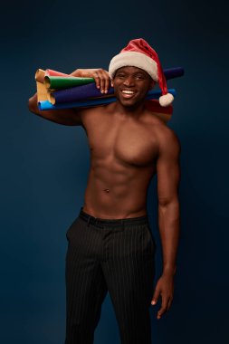 Mutlu, güçlü, siyah mavi üzerine gülümseyen renkli duvar kağıtlarıyla Noel Baba şapkalı Afrikalı Amerikalı adam.
