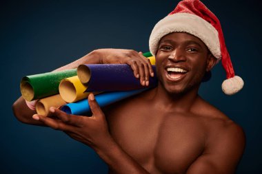 Neşeli ve üstsüz, Noel Baba şapkalı, lacivert renkli duvar kağıtlarıyla gülümseyen Afrikalı Amerikalı.