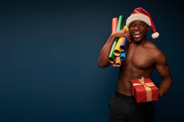 Neşeli ve gömleksiz, Noel Baba şapkalı, koyu mavi renkli hediye kutusu olan Afro-Amerikan adam.