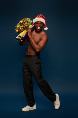 Neşeli, güçlü, Afro-Amerikalı, Noel Baba şapkalı, lacivert arka planda noel dekoru tutan adam.