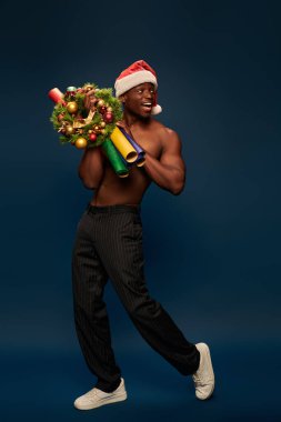Noel Baba şapkalı Afro-Amerikan adam donanma mavisi üzerinde Noel dekoru ile yürüyor.