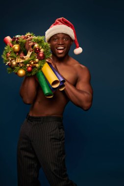Noel Baba şapkalı, koyu mavi üzerinde renkli Noel dekoru olan güçlü, aşırı neşeli Afrikalı Amerikalı adam.