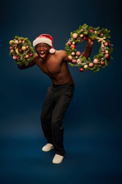 Etkilenmiş ve kaslı Afrikalı Amerikalı Noel Baba şapkalı adam elinde lacivert çelenk tutuyor.