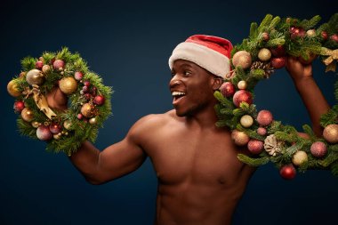 Noel Baba 'lı Afro-Amerikan adam koyu mavi çelenklerle ayakta duruyor.
