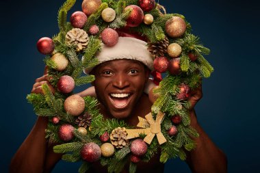 Noel Baba şapkalı Afrikalı Amerikalı adam koyu mavi çelengin içinden kameraya bakıyor.