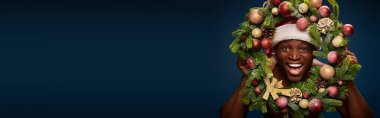 portrait of cheerful american man in santa hat and christmas wreath on dark blue backdrop, banner clipart