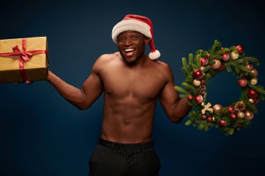 amazed muscular african american with present and christmas wreath looking at camera on dark blue clipart